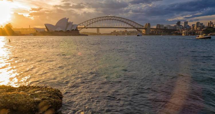 Coucher de soleil sur le port de Sydney avec l'Opéra, le pont du port et les reflets dorés sur l'eau.