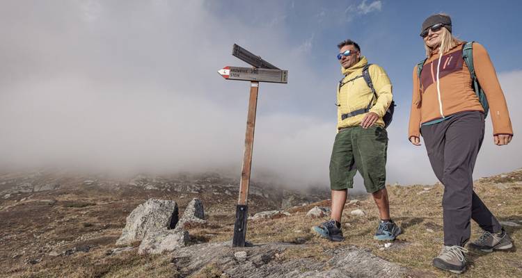 Deux randonneurs marchant sur un sentier de montagne près d'un panneau indicateur brumeux.