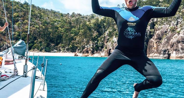 Un viajero vestido con traje de neopreno salta emocionado desde un velero hacia las aguas turquesas de Whitsunday enmarcadas por acantilados boscosos.