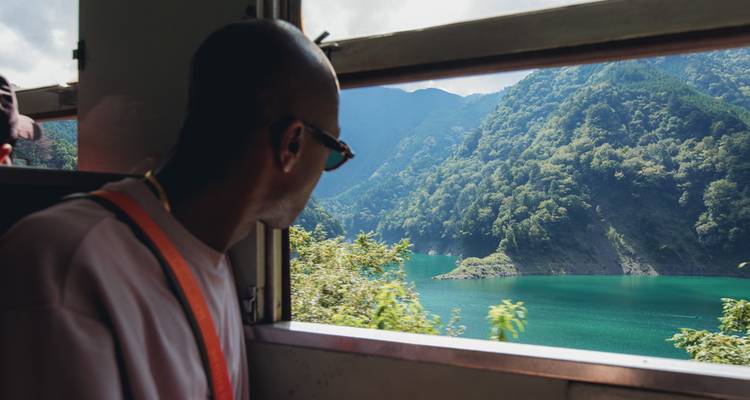 A man gazing out of a train window at lush greenery and a lake.