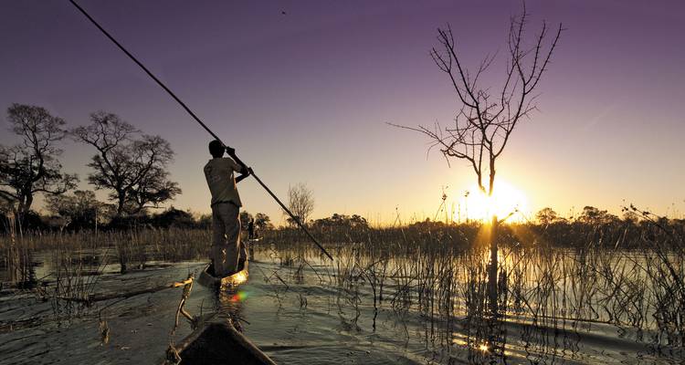 Een persoon die een mokoro met een vaarboom door een kalme waterweg voortduwt bij zonsondergang.