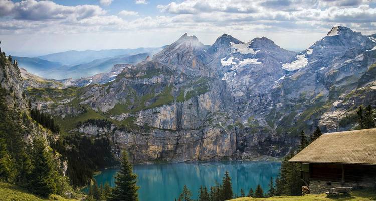 Mountain lake surrounded by cliffs and forest, with a cabin in the foreground.