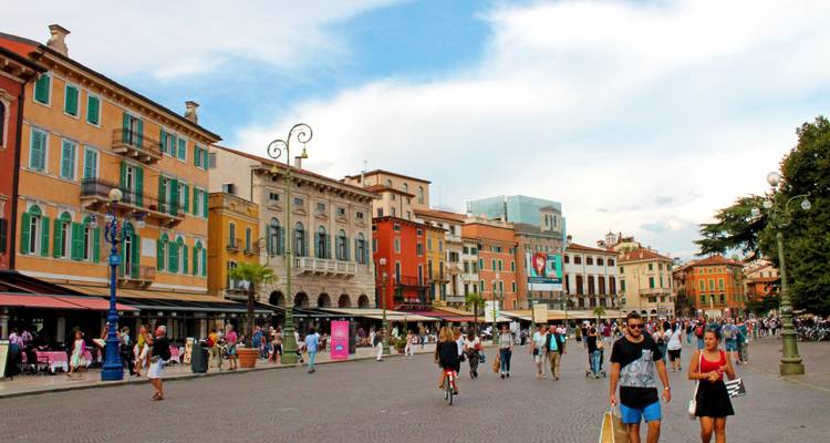 Plaza peatonal bulliciosa bordeada de edificios coloridos, cafés y compradores disfrutando de una tarde soleada.