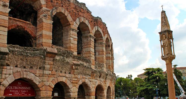 Primer plano de los arcos de piedra antigua de la Arena de Verona junto a una columna alta coronada con una estatua contra un cielo brillante.