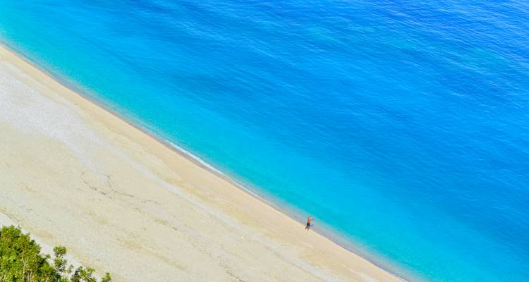 Persoon die langs een levendig strand loopt met helder blauw water.