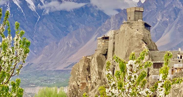 Het historische Baltit Fort kleeft aan een rotsige uitloper met bloeiende bomen op de voorgrond en dramatische Karakoram-toppen erachter