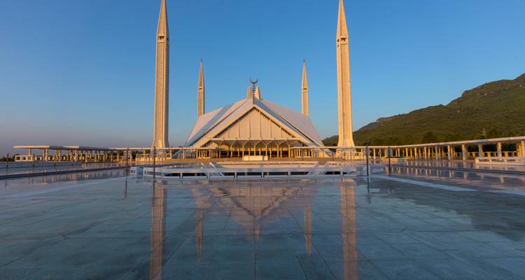 Vue sereine de l'aube de la Mosquée Faisal se reflétant sur la grande cour d'avant-plan en marbre avec un ciel bleu au-dessus.
