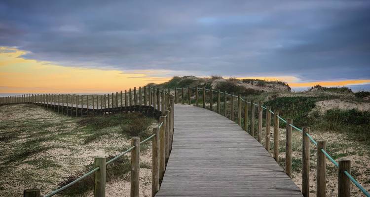 Houten promenade die zich slingerend over kustduinen uitstrekt bij zonsondergang onder stemmige wolken.