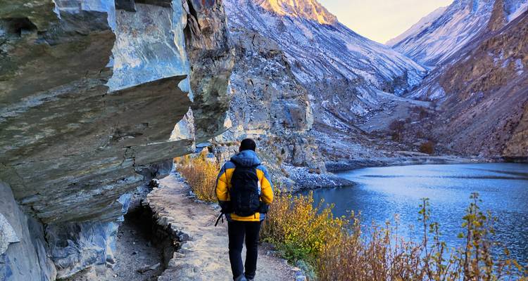 Mochilero con chaqueta amarilla camina por un sendero rocoso junto al lago bajo montañas cubiertas de nieve.