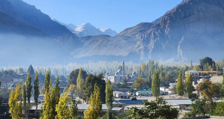 Pueblo del valle lleno de niebla con mezquita de cúpula azul enclavado entre álamos altos y picos rocosos imponentes.