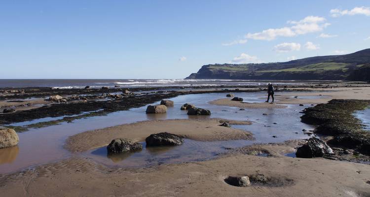 Beach with rocks and a person walking, hills in the background.