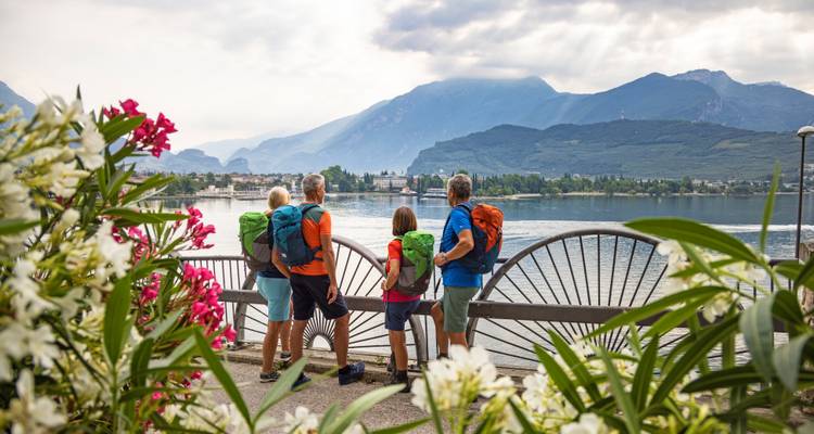 Four hikers with backpacks admire a sparkling lake framed by mountains and flowers.