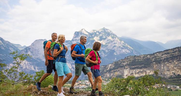 Four enthusiastic hikers trek along a mountain ridge trail with jagged peaks behind.