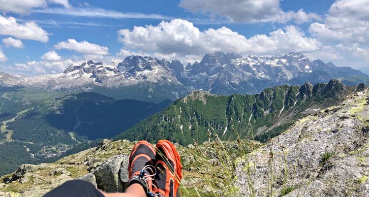A hiker’s shoes rest on a rocky ledge overlooking dramatic snow-capped Dolomite peaks and deep green valleys