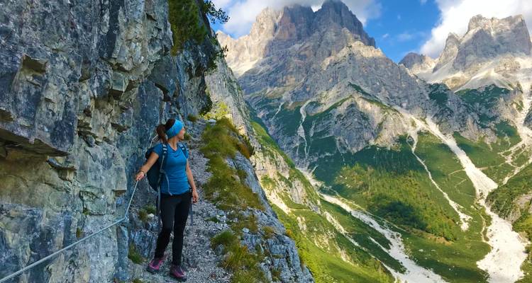Female hiker grips a cable while traversing a narrow mountain path beneath towering Dolomite spires