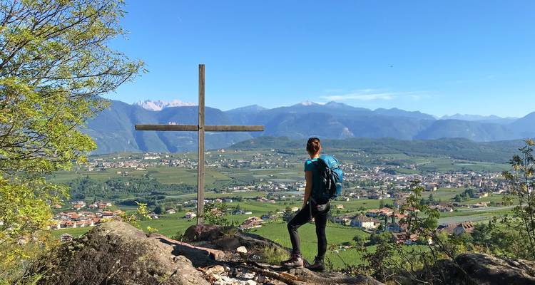 Un viajero se encuentra junto a una cruz de madera con vista a un amplio valle verde respaldado por montañas distantes