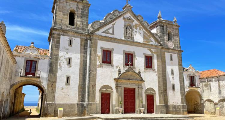 Église-forteresse blanche patinée avec une façade ornée et des portes rouges se dresse contre un ciel portugais d'un bleu éclatant.
