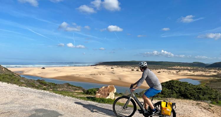 Un cycliste regarde un vaste lagon sablonneux et des dunes ondulantes sous un ciel bleu éclatant strié de traînées de condensation.