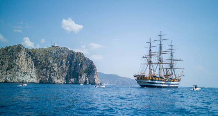 Grand voilier naviguant près d'une île rocheuse sous un ciel bleu.