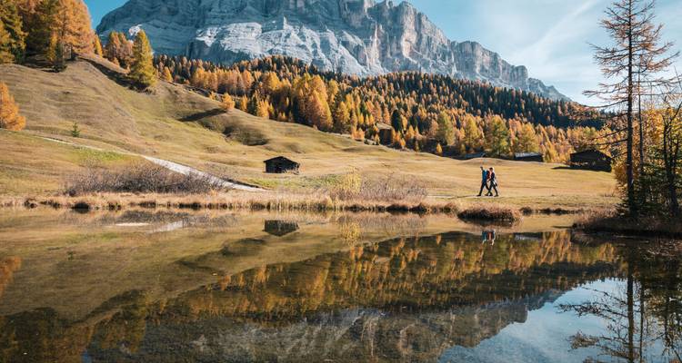 Dos personas caminando junto a un lago que refleja los vibrantes árboles otoñales y las montañas.