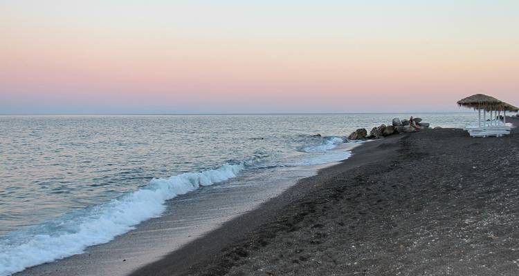 Ruhige Strandszene bei Sonnenuntergang mit dunklem Sand und sanften Wellen.