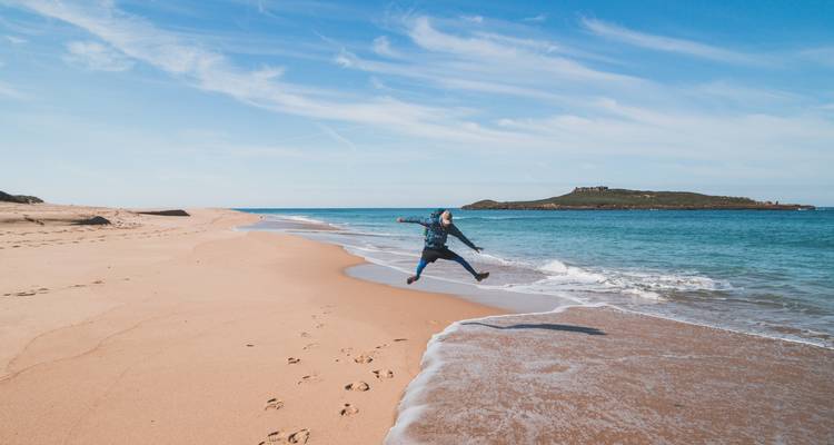 Person springt an einem sandigen Strand mit klarem blauem Wasser.