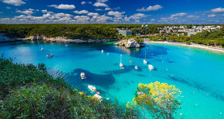 Baie bleu-aqua parsemée de voiliers et bordée de forêts de pins et de plages de sable blanc sous un ciel éclatant.