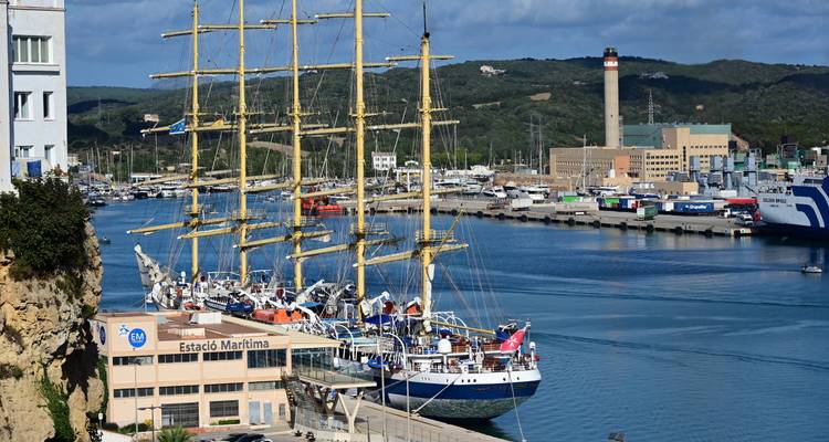 Navire à voiles à mâts élancés amarré dans un port achalandé avec des eaux turquoise et des bâtiments portuaires industriels en arrière-plan.