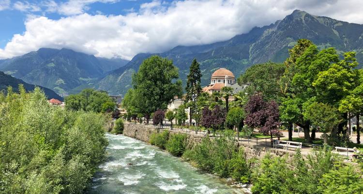 Río alpino de corriente rápida bordeado de árboles con el Kurhaus de cúpula de Merano y altos picos detrás.