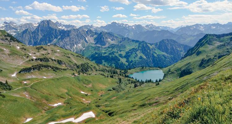 Lago alpino turquesa situado entre prados verdes ondulantes y picos dentados cubiertos de nieve.