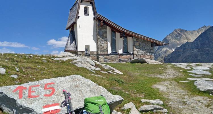 Pequeña capilla de piedra encaramada en una colina alpina rocosa bajo un cielo despejado.