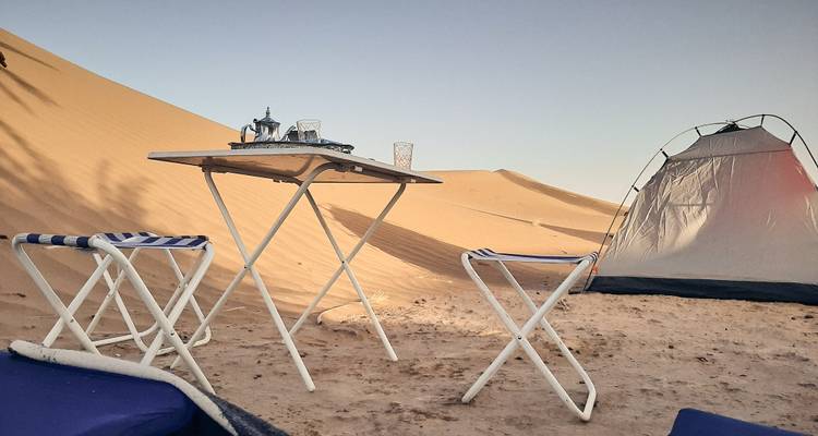 Desert camping setup with a table, chairs, and tent on sand dunes.