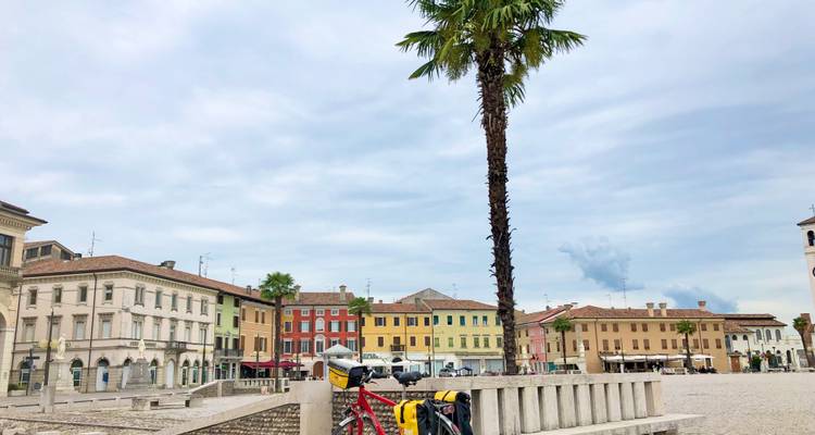 Kleurrijke gevels van een Italiaans stadsplein met een hoge palmboom en toerfietsen beladen met fietstassen op de voorgrond.