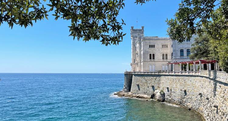 Kasteel Miramare gelegen op een stenen zeedijk boven de diepblauwe Adriatische Zee, omlijst door groene takken onder een heldere hemel.