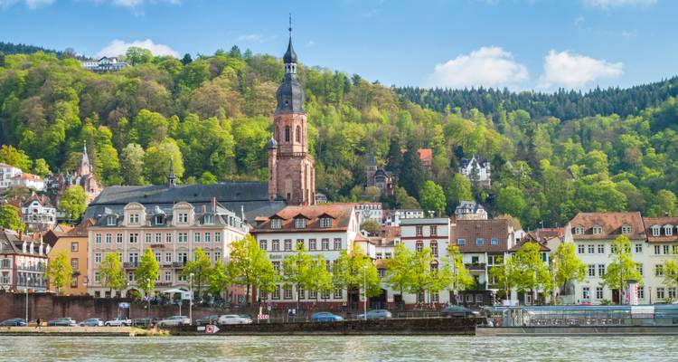 Gebäude entlang des Flusses mit einem Kirchturm und Hügeln.