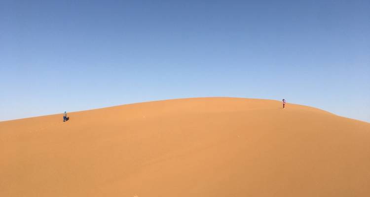 Deux minuscules silhouettes gravissant une dune isolée et imposante sous un immense ciel bleu.