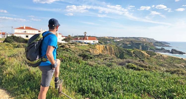 Ein männlicher Wanderer macht eine Pause auf einem Küstenweg mit Blick auf weißgedeckte Häuser, Klippen und den Atlantischen Ozean.