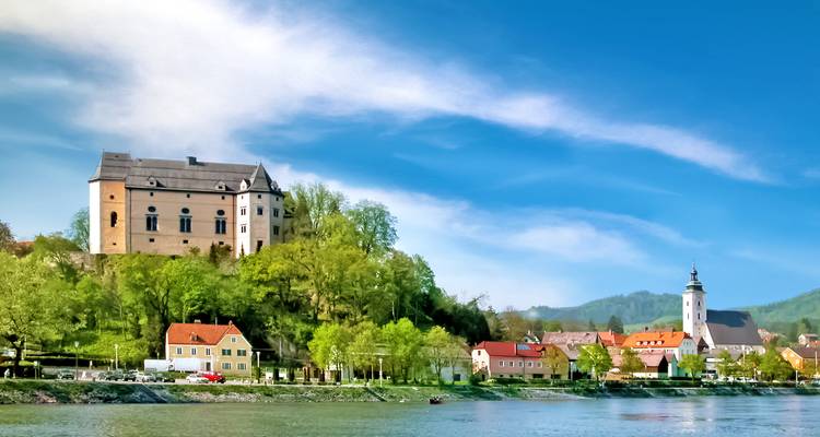 Un castillo majestuoso corona una colina boscosa sobre un tranquilo pueblo ribereño con una torre de iglesia, todo bajo un cielo azul vívido.