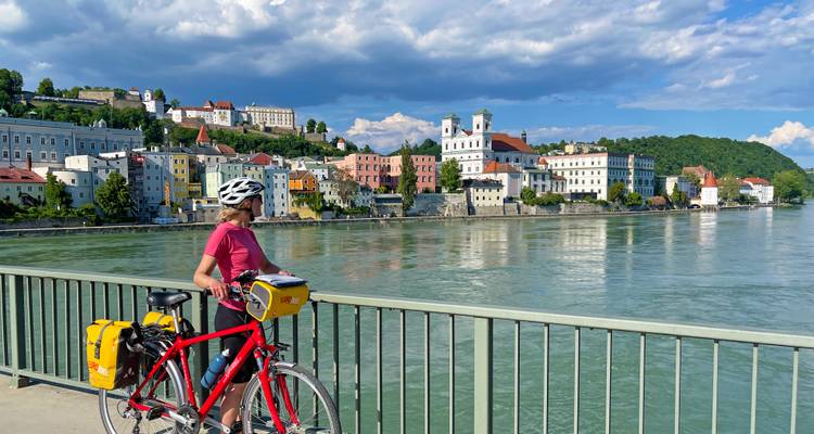 Un ciclista solitario se encuentra en un puente con vista a coloridos edificios ribereños y torres de iglesias reflejadas en el agua.