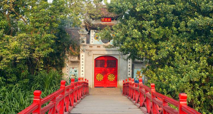 Pont rouge orné menant à un temple traditionnel entouré d'arbres.