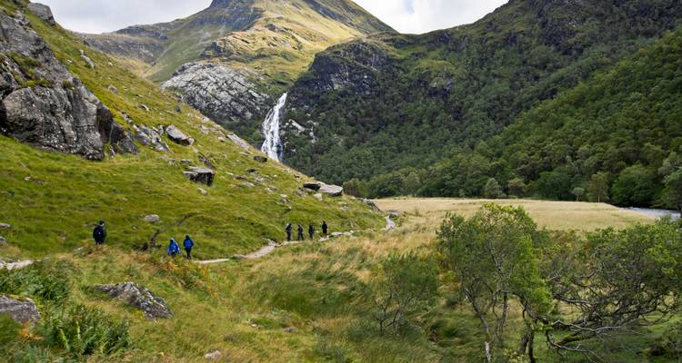 Hikers walking along a lush green valley with a waterfall in the distance.