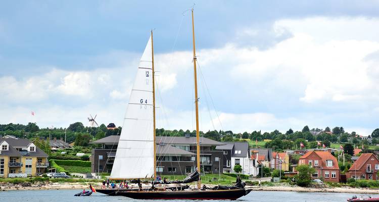 Sailboat on a waterway with houses and greenery in the background.