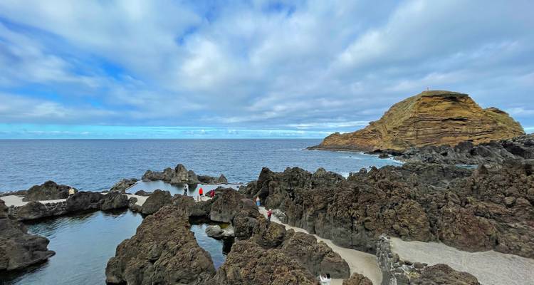 Piscines naturelles de roche volcanique avec visiteurs et promontoire escarpé sous un ciel partiellement nuageux.