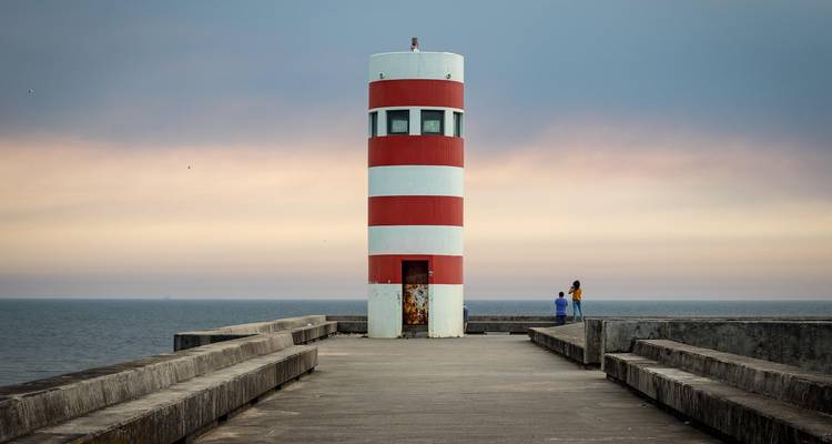 Phare à rayures rouges et blanches au coucher du soleil avec deux personnes au loin.