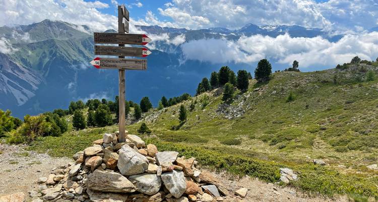 Alpenpadsignaalpost bovenop een rotsachtige steenhoop met panoramische bergkammen en wolkenlagen erachter.