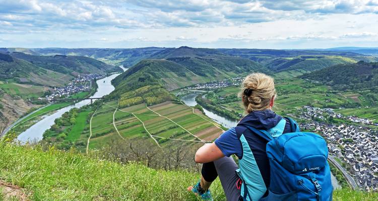 Vrouwelijke wandelaarster met blauwe rugzak zit op een grasrijke klif en kijkt uit over slingerende wijngaarden in het Moezel-dal.