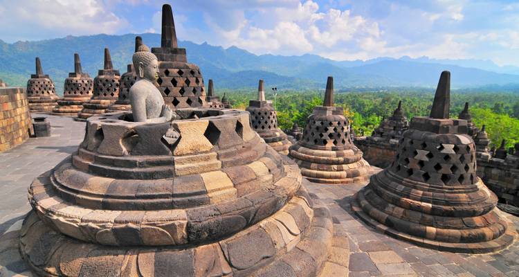 Statues and stupas on a temple platform with a forest and mountain backdrop.