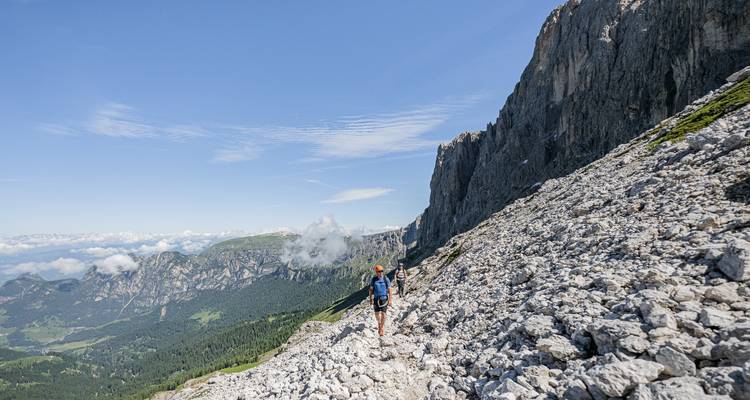 Un excursionista atraviesa una ladera montañosa rocosa bajo espectaculares acantilados de piedra caliza con amplias vistas alpinas bajo un cielo azul despejado.