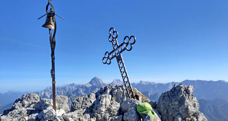 Escena de cumbre con campanario de hierro y cruz metálica elevándose desde picos calizos escarpados bajo un cielo azul despejado.