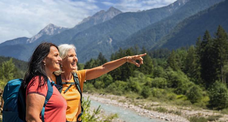 Dos excursionistas mayores sonríen y señalan hacia picos alpinos distantes junto a un río cristalino.